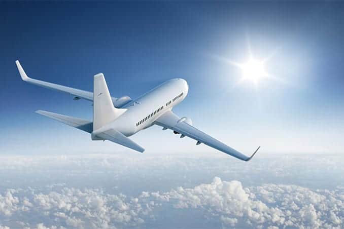 A white commercial airplane in flight above the clouds with the sun shining brightly in the clear blue sky.