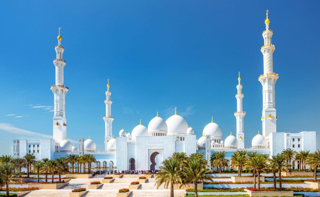 A panoramic view of the Sheikh Zayed Grand Mosque in Abu Dhabi, featuring its magnificent white domes and minarets against a clear blue sky. The foreground includes symmetrical gardens and palm trees, while the mosque's intricate architectural details are highlighted; it's a must-see on Dubai tour packages from USA.