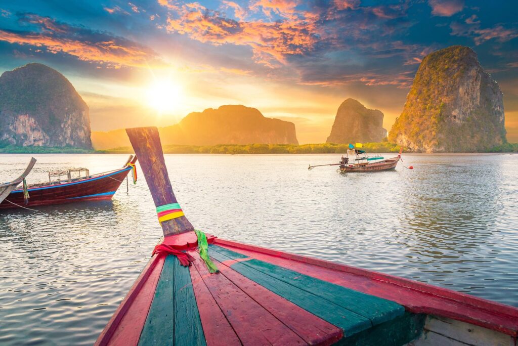 A serene image of a vibrant longtail boat on calm waters during sunset, with dramatic cliffs and lush greenery in the background. Another boat is seen in the distance under a sky painted with warm colors of the setting sun, evoking dreams of Thailand and Phuket tour packages.