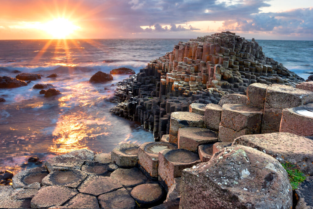 A scenic view of the Giant's Causeway in Northern Ireland at sunset. The hexagonal basalt columns are illuminated by the soft golden rays of the setting sun, with waves gently lapping against the rock formations and a colorful sky in the background—truly a highlight of Ireland escorted tours.