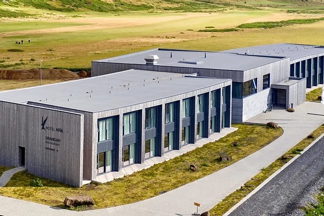 Aerial view of the modern, rectangular "Hotel Kría" with large windows. Nestled in an open, grassy landscape with a paved path to its entrance, it's an ideal retreat for those on an Iceland tour seeking the serene beauty and tranquility perfect for catching glimpses of the Northern Lights.