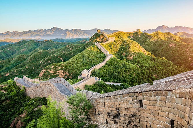 A panoramic view of the Great Wall of China winding through lush green mountains under a clear blue sky. The ancient wall, built with stone and bricks, stretches across ridges and valleys, showcasing its architectural grandeur and historic significance.