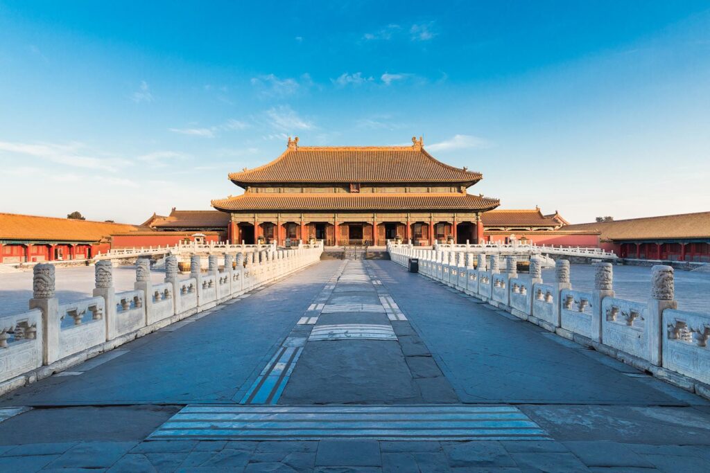 A wide-angle view of an ancient, ornate building in the Forbidden City in Beijing, China. The structure features traditional Chinese architecture with a sloping roof and intricate details. A stone pathway and a series of white stone railings lead to the entrance. The sky is clear and blue.