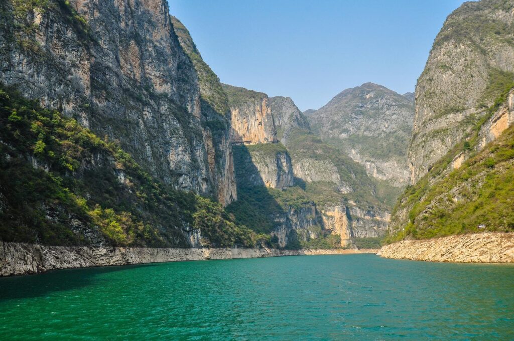 A stunning river scene shows turquoise waters flanked by towering rocky cliffs and lush green vegetation. The cliffs rise dramatically on both sides under a clear blue sky, creating a breathtaking and serene canyon landscape.