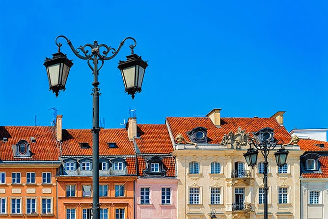 A row of colorful, traditional European buildings with red-tiled roofs under a clear blue sky. Ornate black street lamps are in the foreground, adding a touch of historic charm to this vibrant Prague scene.