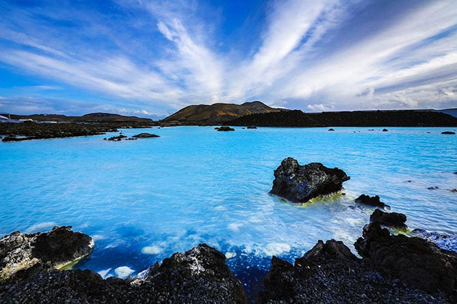 A stunning landscape with a vibrant blue geothermal lagoon surrounded by dark volcanic rocks awaits on our Iceland Escorted Tours. The sky above is filled with dramatic white clouds stretching outward, and distant hills can be seen on the horizon.