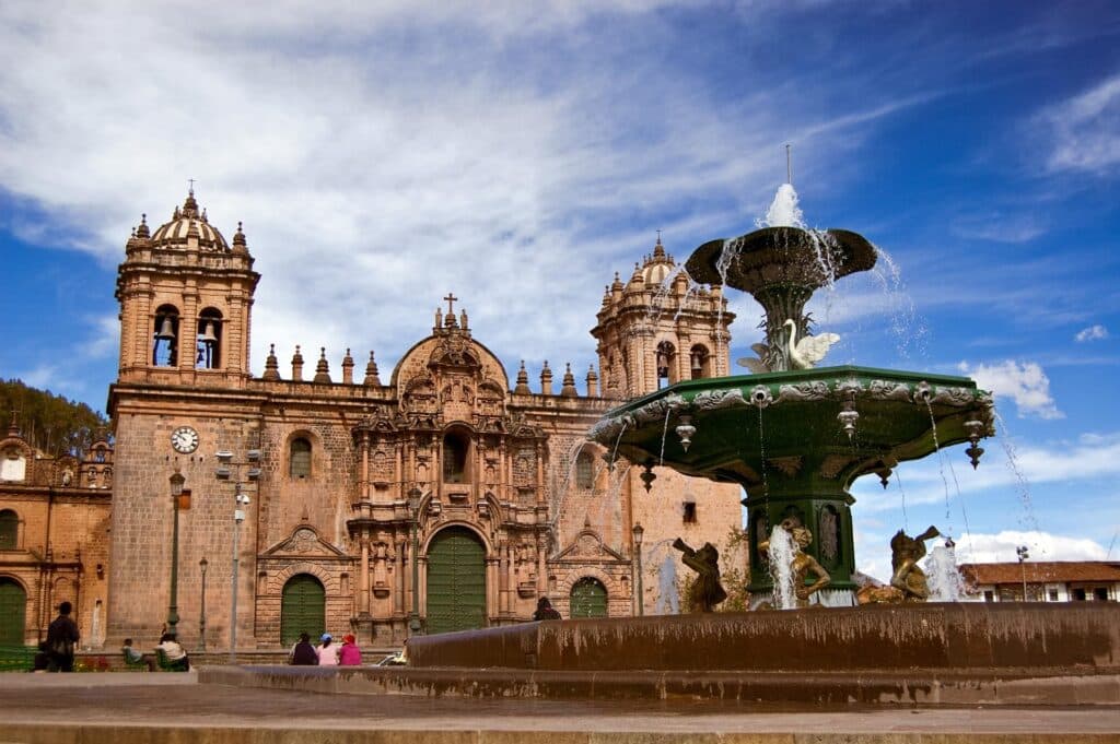 An ornate fountain in a plaza with a historic cathedral in the background, featuring intricate stonework and twin bell towers. A few people sit near the fountain under a partly cloudy sky, reminiscent of scenes from Peru vacation packages with airfare included.