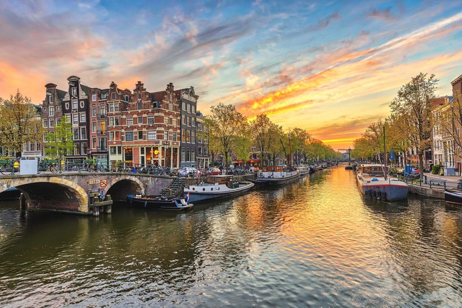 Scenic view of an Amsterdam canal at sunset. Historic buildings line the waterway, with a bridge on the left and boats moored along the sides. Consider river cruise package tours to experience these vibrant orange and pink hues reflecting in the calm water, bordered by trees with autumn foliage.