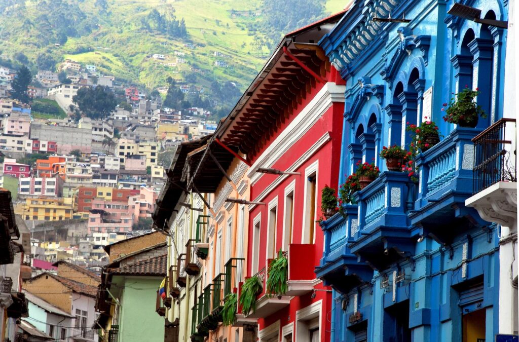 Colorful buildings with balconies line a narrow street in a hilly, densely populated area. The vibrant facades include blue, red, and orange tones, contrasting with the green hills and numerous houses in the background—a perfect stop on your Ecuador and Galapagos Islands tour. Flowers adorn many of the balconies.