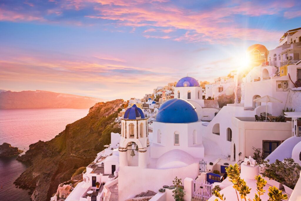 A stunning view of Santorini, Greece at sunset, featuring iconic white-washed buildings with blue domes overlooking the Aegean Sea. The sun casts a warm glow on the architecture, highlighting the cliffside village and vibrant sky with hues of pink and orange during your Spectacular Greece tour.