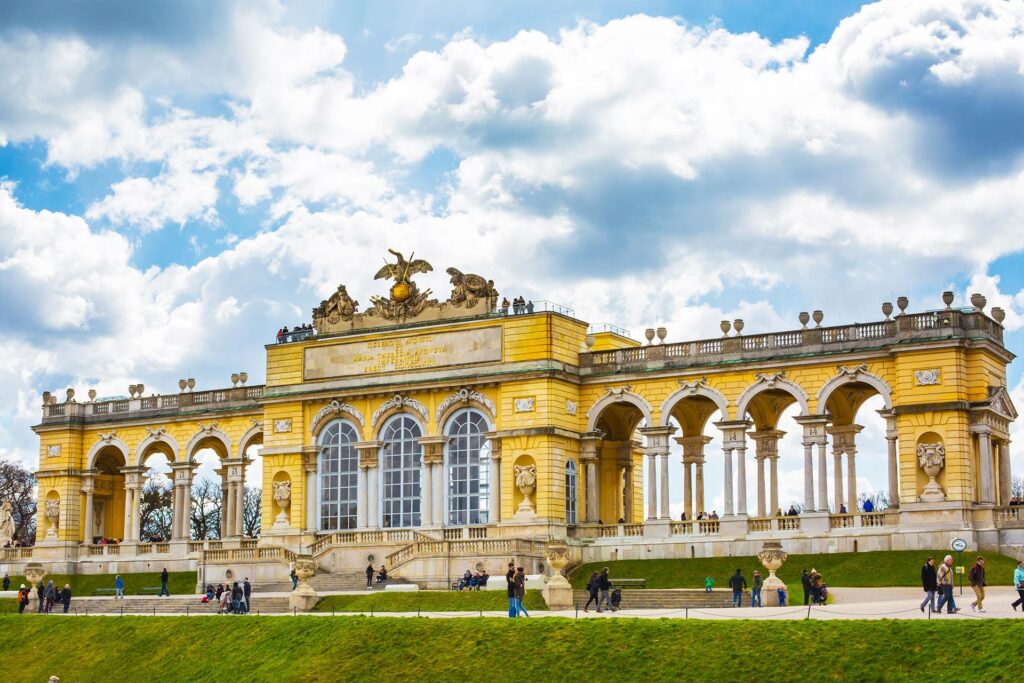 A grand, yellow Baroque-style pavilion with arched windows and columns stands under a partly cloudy sky, reminiscent of those seen in Vienna. People are walking and sitting on the grass in front of the building. Sculptures and a large golden eagle adorn the rooftop, creating an inviting stop on any European tour.