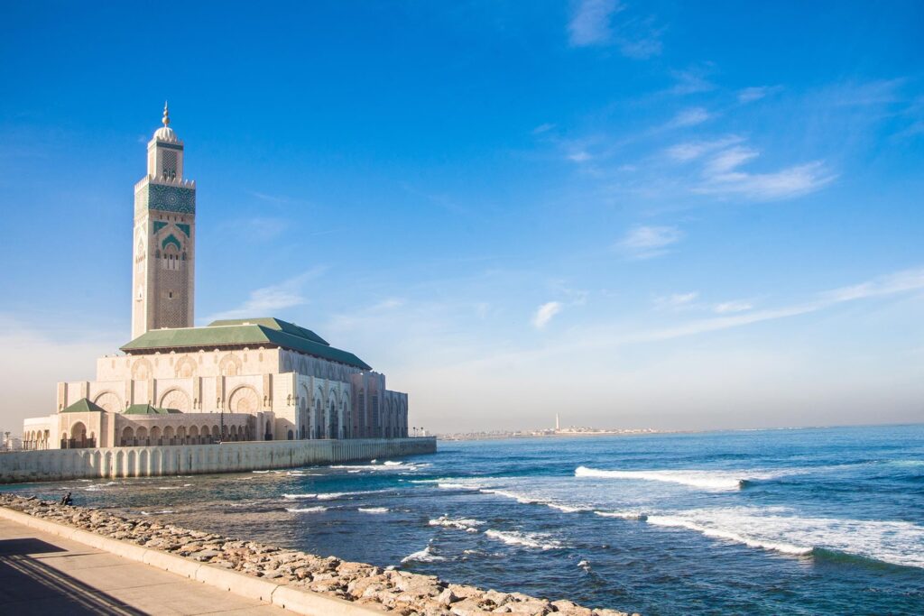 A mosque with a tall minaret stands beside the ocean under a clear blue sky, frequently featured in Morocco Escorted Tours. The building boasts intricate architectural details and arches, while waves gently reach the rocky shore in the foreground. A distant coastline is visible on the horizon.