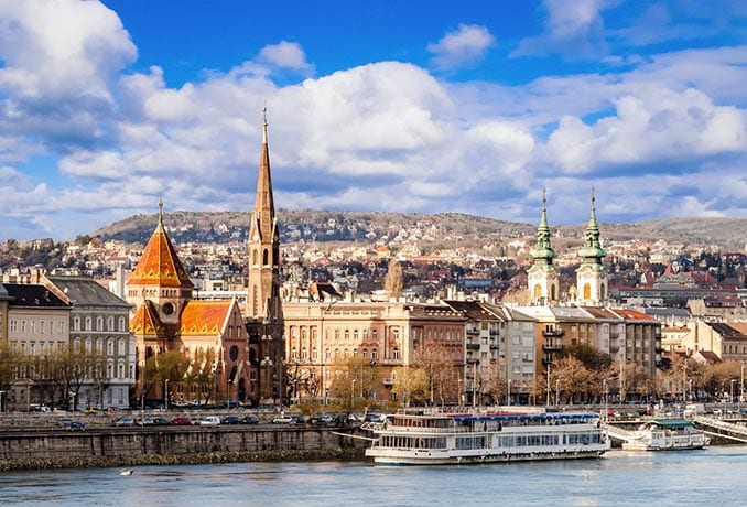 Panoramic view of Budapest, Hungary, showcasing classic European architecture. Prominent features include the pointed tower of the Matthias Church and ships on the Danube River. This Budapest tour reveals a cityscape stretching out with buildings and hills under a partly cloudy sky.