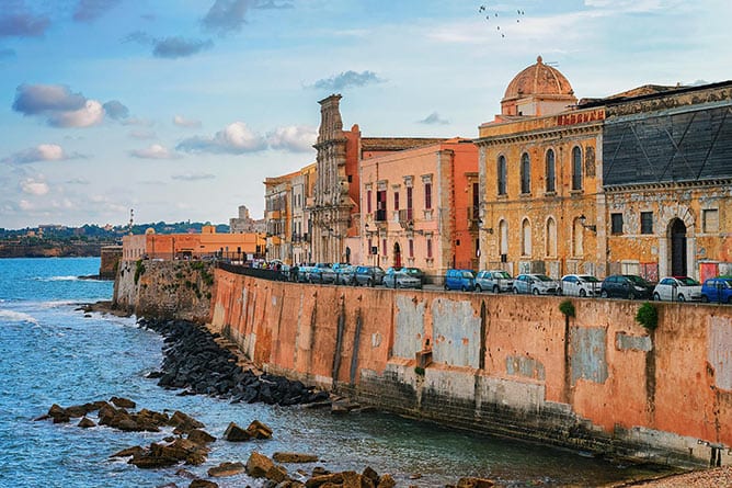 A seaside view of a historic Italian town with a row of colorful buildings along the waterfront. The architecture, showcasing various textures and colors, overlooks a calm, rocky shoreline. Cars are parked along the street, and birds fly overhead—an inviting scene for tours of Sicily enthusiasts.