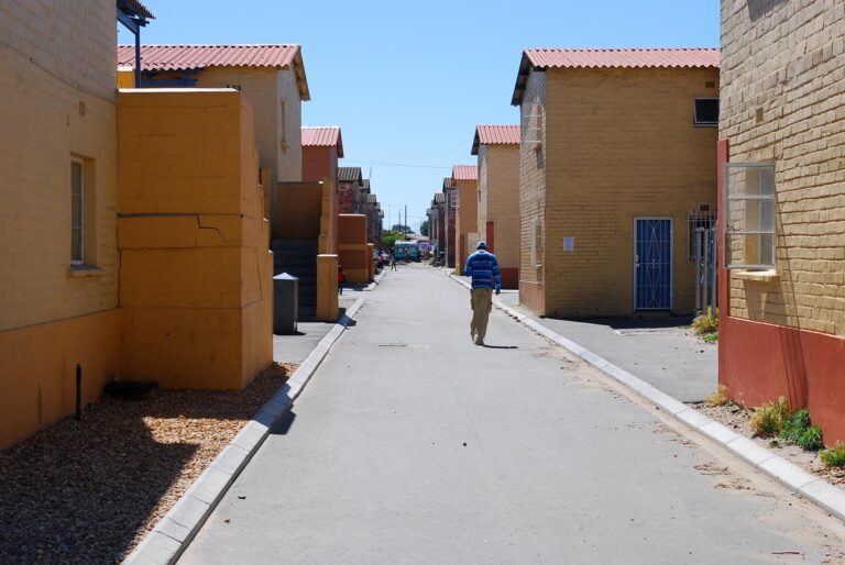 A person strolls down a narrow street lined with tall, yellow, and brown brick buildings on a sunny day, reminiscing about their recent South Africa tour.