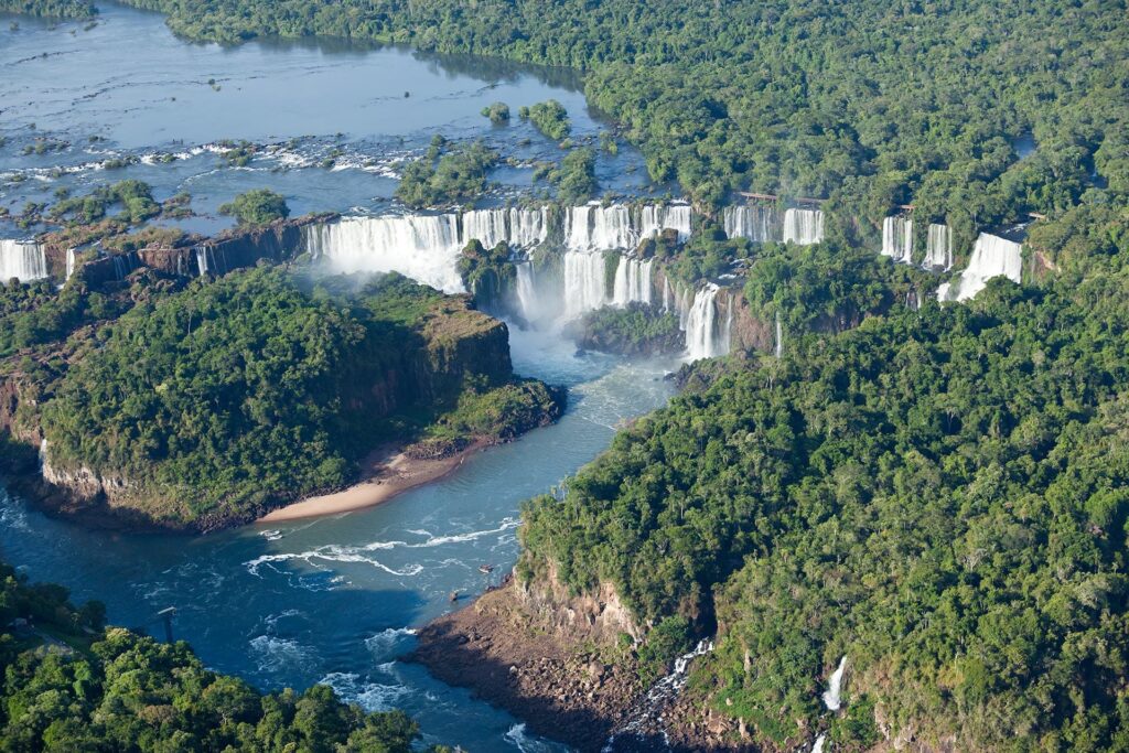 Aerial view of Iguazu Falls, one of the highlights of South America, where numerous waterfalls cascade down cliffs surrounded by lush green rainforest. The extensive water flow creates misty clouds, and the river below winds through dense vegetation. The sky above is clear and bright.