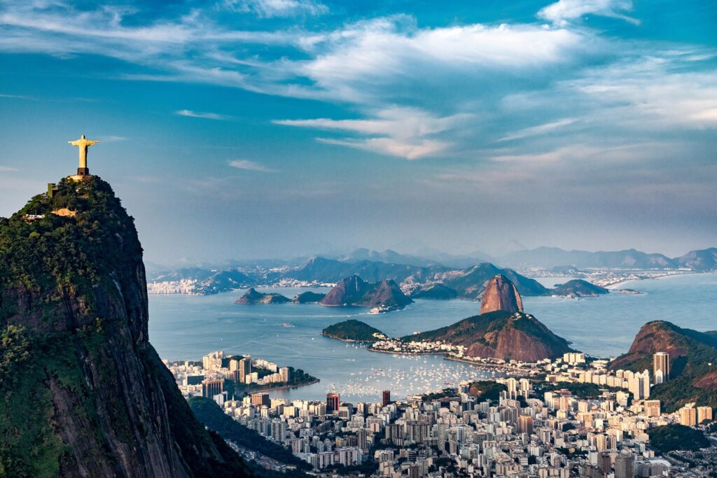 Aerial view of Rio de Janeiro, Brazil, highlighting the Christ the Redeemer statue atop Corcovado Mountain, overlooking the South American cityscape, coastal waters, and surrounding mountain peaks under a partly cloudy sky.