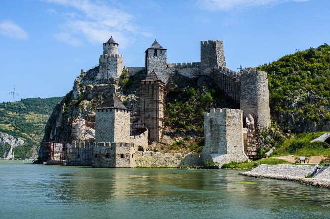 A medieval stone fortress with multiple towers is situated on a hillside next to a body of water. The structure is partially under renovation, as scaffolding can be seen on some of its sections. The sky is clear, and surrounding the fortress is lush greenery.