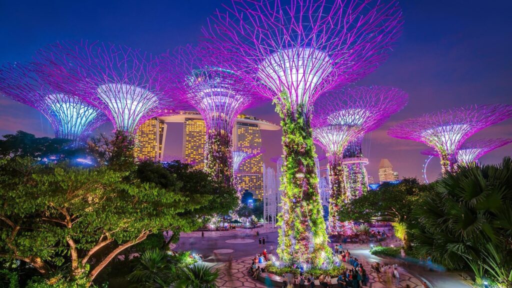 A vibrant nighttime scene of futuristic tree-like structures illuminated in pink and purple, located in Gardens by the Bay, Singapore. The Marina Bay Sands hotel stands majestically in the background against a dusky sky, reminiscent of cityscapes seen in Hong Kong, surrounded by lush greenery and visitors.