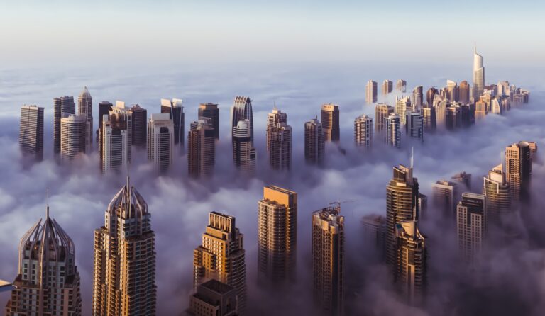 Aerial view of a city skyline with numerous skyscrapers emerging through a thick blanket of fog. The buildings vary in architectural style and height, creating a dramatic and surreal scene as they rise above the mist. The sky is clear and light blue, much like the vistas on Dubai tour packages from the USA.