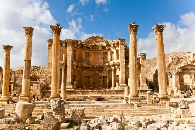 Ancient Roman ruins featuring tall, stone columns and the remnants of a stone structure under a bright blue sky with scattered clouds. Rubble and broken columns are scattered in the foreground, and the scene reflects a historical architectural marvel.