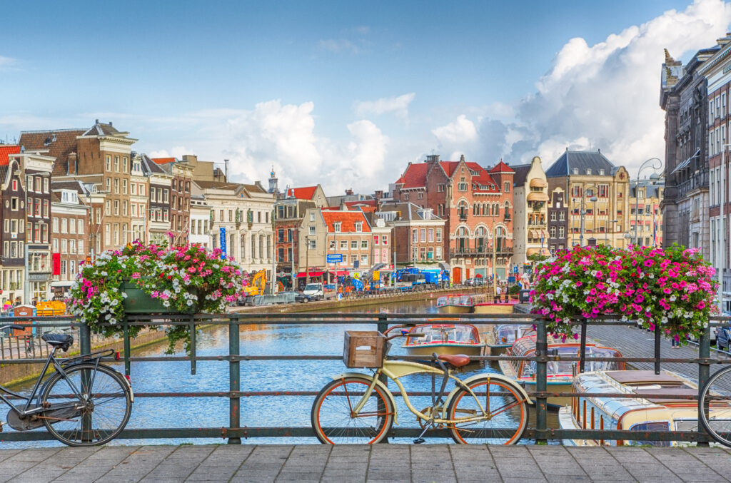 Scenic view of an Amsterdam canal with historic buildings, boats, and a bridge adorned with vibrant flower baskets and a parked bicycle. The sky is partly cloudy, adding to the picturesque and lively atmosphere of the location.