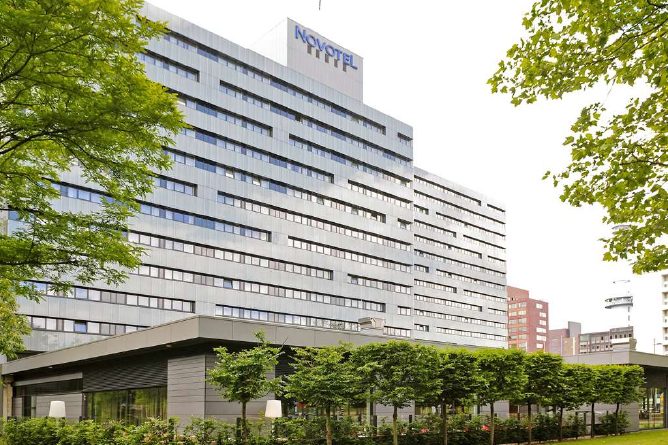 A modern high-rise building with the "Novotel" logo on top stands amidst greenery. The structure features large windows and a gray exterior. In the foreground are trimmed trees and neatly landscaped plants, with taller buildings visible in the background.