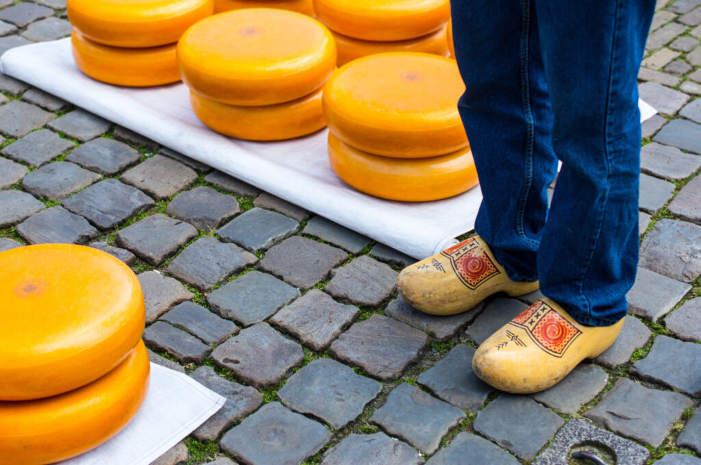 A traveler in yellow wooden clogs stands next to wheels of orange cheese arranged on a white blanket. The cobblestone street scene, bathed in the fresh air of springtime, features blue jeans and a quintessentially Dutch view, evoking the charm of the Netherlands.