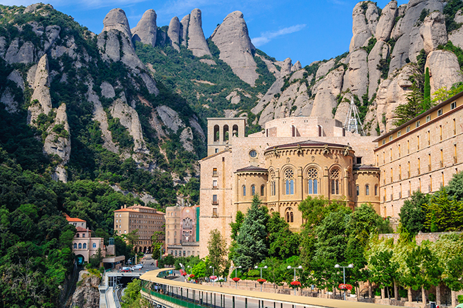 A historic monastery complex built against the backdrop of dramatic, jagged mountains. The sunlit buildings include an ornate church with large windows and towers. Lush green trees and pathways with a few people are seen in the foreground.