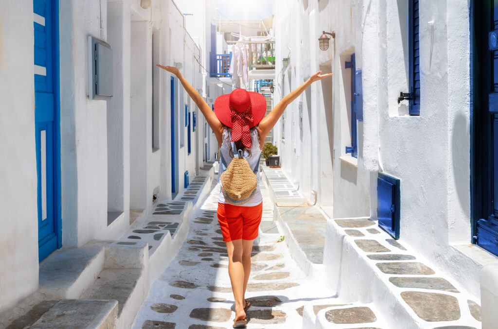 A person wearing a red hat, red shorts, and a backpack joyfully walks down a narrow, white-walled alley with blue doors and windows. The alley is bathed in bright sunlight, evoking a sense of summer and adventure.