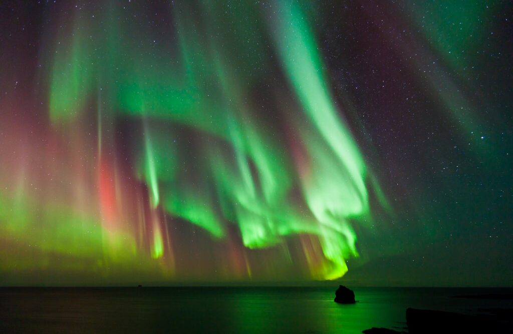 A dazzling display of the Northern Lights featuring vibrant green and red hues illuminates the night sky over a tranquil body of water with a silhouette of a small rock formation or islet in the distance. Experience this magical phenomenon on an Iceland tour with Northern Lights, where stars faintly sparkle in the background.