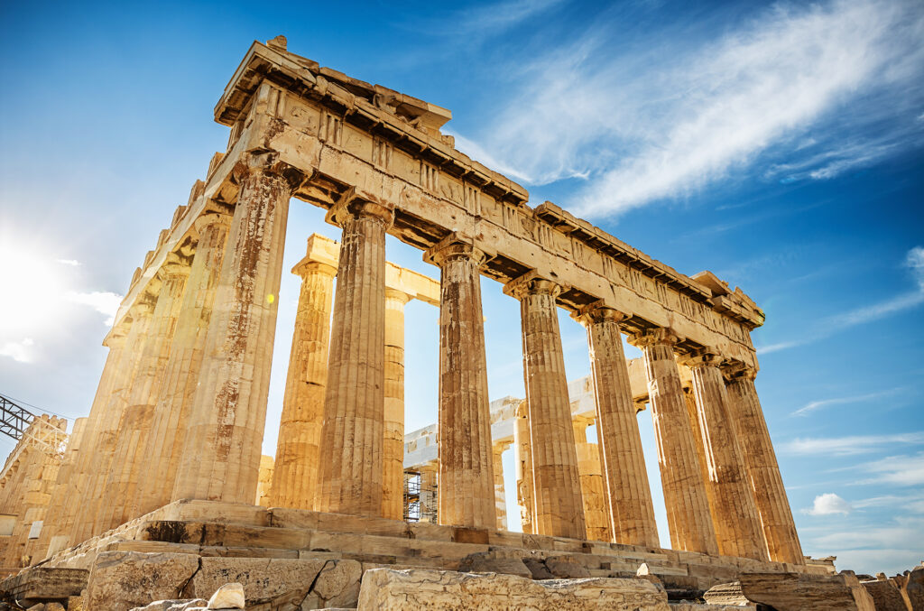 The image features the Parthenon, an ancient Greek temple on the Athenian Acropolis, bathed in Mediterranean sunlight. It showcases its monumental Doric columns and detailed architectural elements against a vibrant blue sky with scattered clouds—a perfect sight for any Three Continents cruise.