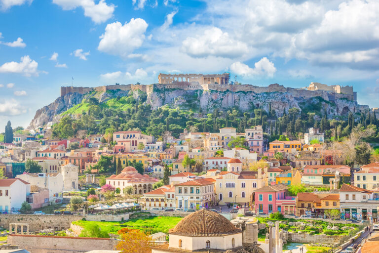 A vibrant cityscape with colorful buildings, lush greenery, and the ancient Acropolis with the Parthenon prominently atop a rocky hill under a partly cloudy sky in Athens, Greece, beckons travelers embarking on a Mediterranean Cruise across Three Continents.