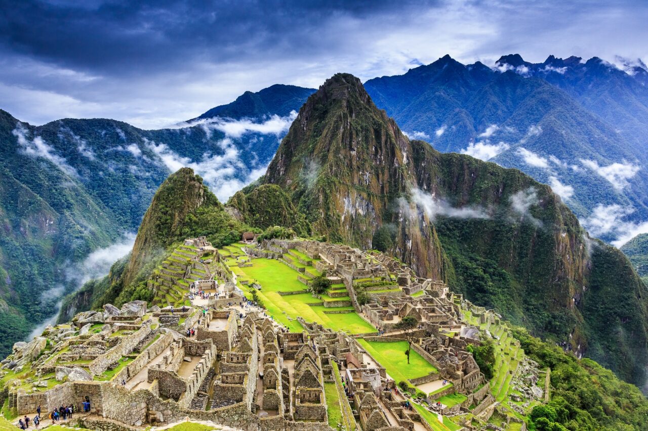 A panoramic view of Machu Picchu, an ancient Incan city set high in the Andes Mountains with lush greenery and mist-covered peaks in the background. The stone ruins are surrounded by terraced fields and dramatic mountainous scenery under a cloudy sky.