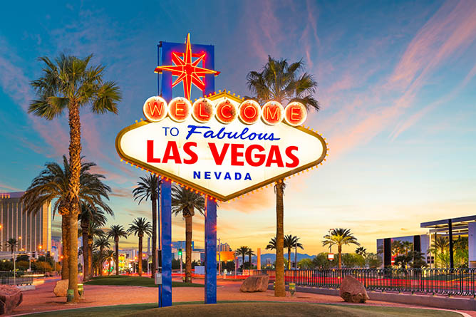 A brightly lit "Welcome to Fabulous Las Vegas Nevada" sign stands against a backdrop of palm trees and a vibrant sunset sky. Buildings and a clear sky with scattered clouds are visible in the distance. The sign is iconic with red, white, and blue colors.