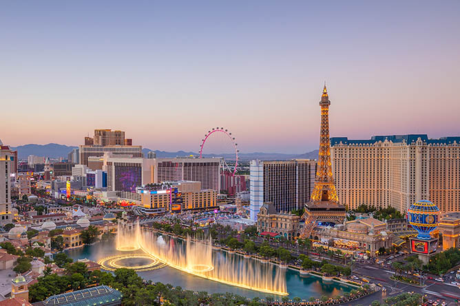 A panoramic view of the Las Vegas Strip at dusk, featuring the illuminated fountains of the Bellagio, the replica Eiffel Tower, and the High Roller Ferris wheel in the background, set against a backdrop of hotels and casinos.