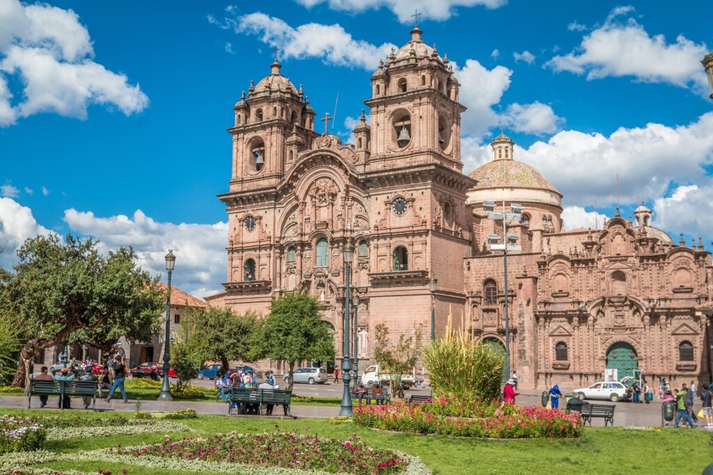 A historic cathedral with ornate architecture stands under a bright blue sky with scattered clouds. In the foreground, a park with green grass, flowers, and people walking or sitting on benches adds to the vibrant scene, reminiscent of a stop on a Galapagos Escorted Tour.