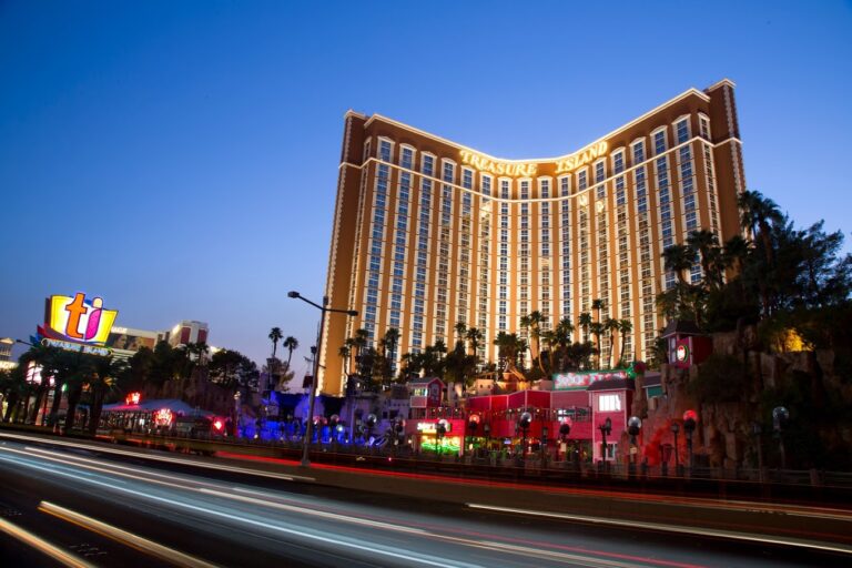 The image shows the Treasure Island Hotel and Casino in Las Vegas illuminated at dusk. The building features prominent neon signage and is surrounded by palm trees. Bright lights from passing vehicles create streaks on the road in the foreground, adding to the sense of adventure in this vibrant Southwest destination.