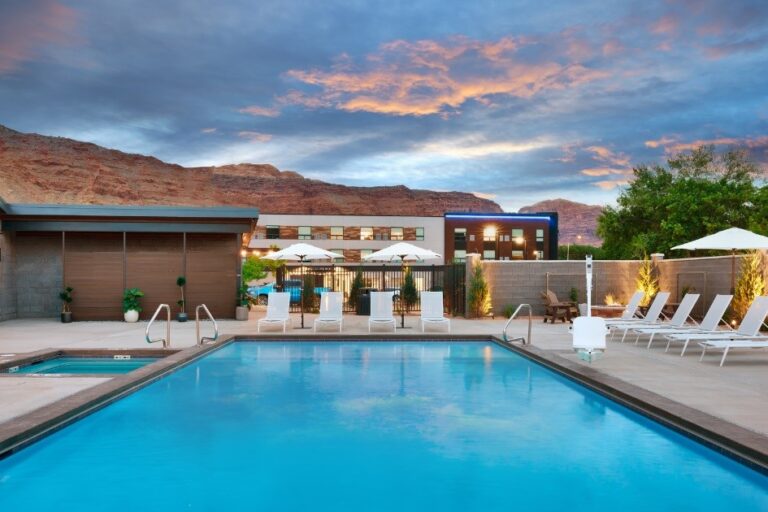 A tranquil outdoor swimming pool with several white lounge chairs, umbrellas, and a hot tub nearby. The area is surrounded by a stone fence and features desert mountains in the background, evoking the spirit of Southwest adventure beneath a dramatic evening sky of blue and orange hues.