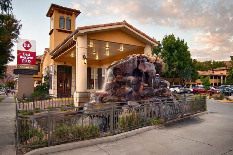 A two-story Best Western hotel with a beige exterior and a stone façade around the entrance. In the foreground, there's a large rock waterfall feature surrounded by a metal fence. The hotel signage is visible to the left, and several cars are parked to the right, ready for your Southwest adventure near National Parks.