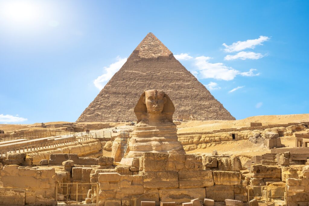 The image shows the Great Sphinx and the Pyramid of Khafre under a clear blue sky. The Sphinx, a limestone statue with a lion's body and a human head, stands in front of the ancient pyramid, surrounded by ruins and desert terrain—a timeless scene anticipating your 2024 departures.