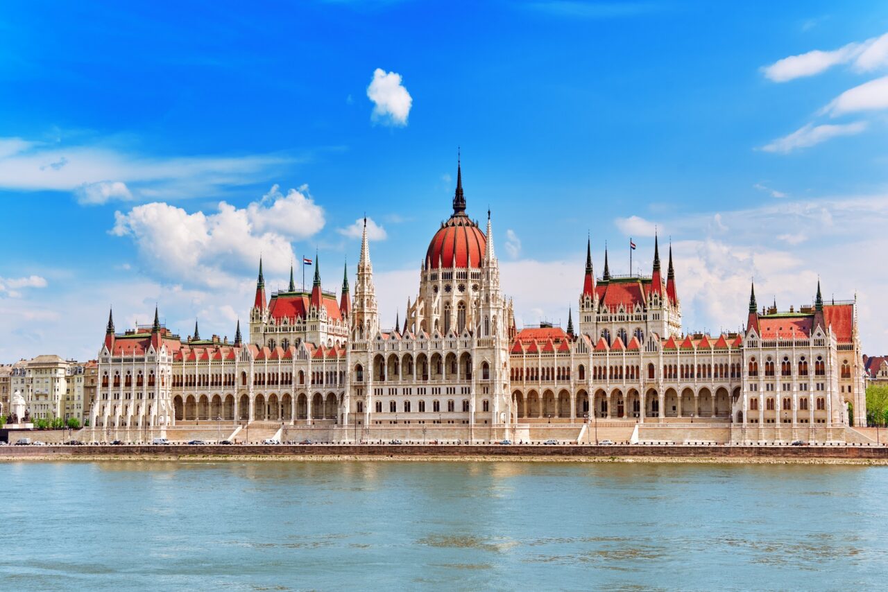 The image shows the Hungarian Parliament Building in Budapest, located alongside the Danube River. The building features a central dome flanked by symmetrical spires and towers, with a clear blue sky and some scattered clouds in the background, evoking dreams of 2024 departures to this stunning landmark.