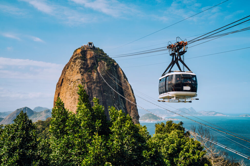 A cable car ascends towards the rocky peak of Sugarloaf Mountain, one of the South American highlights, surrounded by lush greenery and overlooking a scenic coastal view with calm blue waters under a clear blue sky.