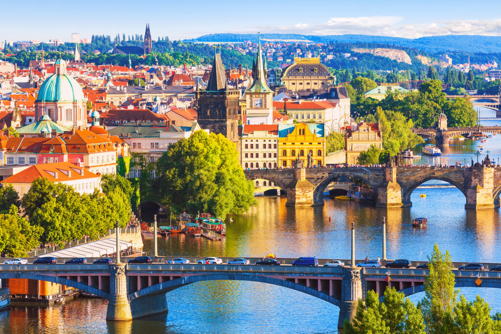 A vibrant cityscape of Prague featuring historic buildings, bridges, and the Vltava River. The Charles Bridge is prominently shown with its statues and towers, while modern and older architecture blend seamlessly under a bright blue sky—an ideal starting point for the Prague-Vienna-Budapest tour.