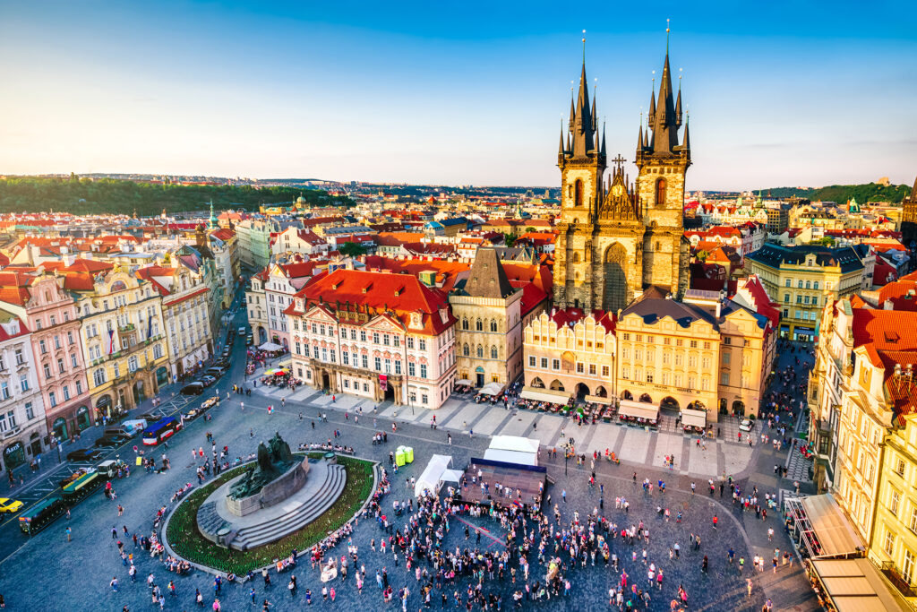 Aerial view of Old Town Square in Prague bustling with people. The square features the Gothic Church of Our Lady before Týn, historic buildings with vibrant red rooftops, and an equestrian statue. The impressive cityscape extends into the background beneath a clear blue sky—perfect for your Prague Vienna Budapest tour.