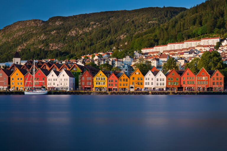 Colorful wooden buildings reminiscent of a Scandinavian explorer's journey line the waterfront in front of a forested hillside. Painted in vibrant reds, yellows, and whites, they reflect in the calm water. A boat is docked on the left, and residential houses are visible on the hillside in the background.