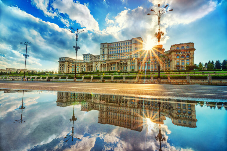 A grand, ornate building under a partly cloudy sky with the sun shining brightly behind it. The building and sun are reflected in a large puddle on the pavement, creating a mirror image. Tall street lamps line the street in front of the structure.