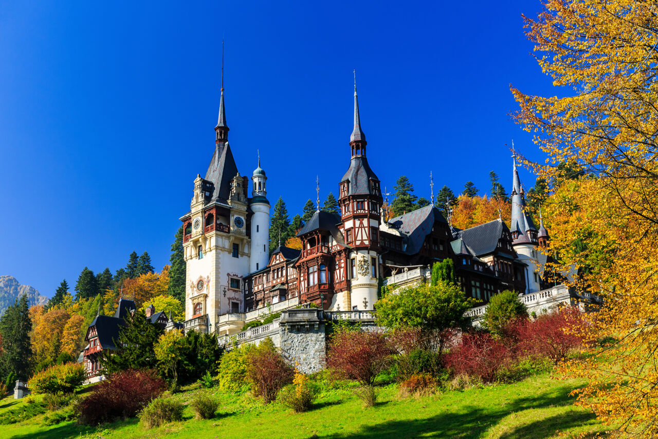 Peleș Castle in Romania, surrounded by lush green grass and trees with autumn foliage. The sky is clear and bright blue, highlighting the castle's intricate architecture with multiple towers and detailed designs. The scene is vibrant and picturesque.