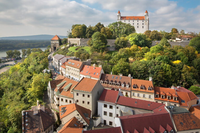 A scenic view of Bratislava Castle perched atop a hill, surrounded by lush greenery, with a town of colorful buildings and red-tiled roofs in the foreground. The sky is partly cloudy, allowing sunlight to subtly illuminate the landscape.