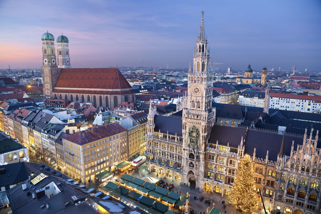 Aerial view of Marienplatz in Munich, Germany, during the evening. The square is lit with festive holiday lights, a large decorated Christmas tree, and numerous Christmas markets. The Neue Rathaus and the twin towers of the Frauenkirche are prominent in the background.